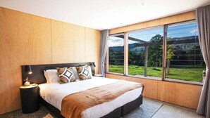 A modern bedroom with a large bed, a nightstand, and a view of a green landscape through large windows. - Jacaranda at Barranca - Luxury Villa (Kangaroo Valley)