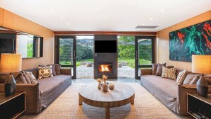 A modern living room with a fireplace, two sofas, a coffee table, and large windows offering a view of the outdoors. - Jacaranda at Barranca - Luxury Villa (Kangaroo Valley)