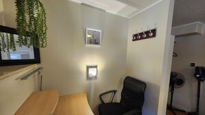 A room with a wooden desk, a black office chair, a hanging plant, a framed picture, a wall-mounted light, and a ceiling fan.