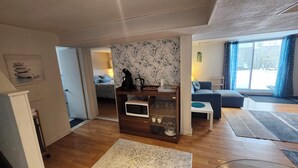 A modern living room with a wooden floor, a coffee table, a sofa, a wall-mounted shelf with a kettle, and a view through a large window.