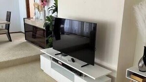A modern living room with a flat-screen TV on a white entertainment unit, a coffee table, and a plant. - A three bedroom house with 2 bathrooms (Gresham)