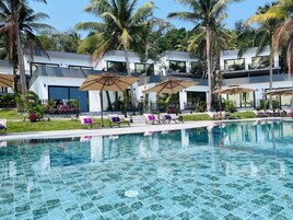 Una piscina con piso de azulejos, rodeada de sillas de descanso y sombrillas, con palmeras y un edificio moderno al fondo.