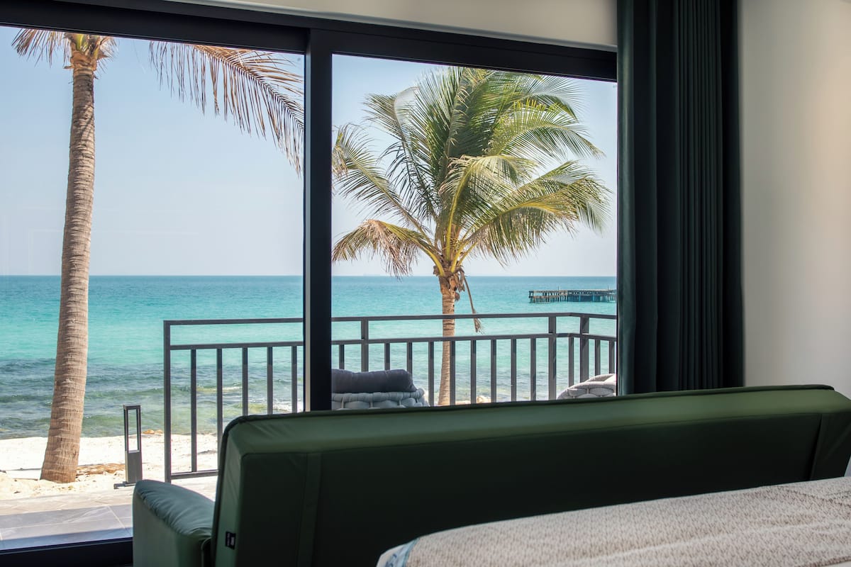 A room with a large window overlooking a beach, a palm tree, and a view of the ocean.