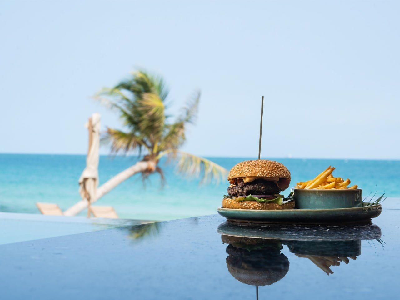 Un plato con una hamburguesa y papas fritas sobre una superficie reflectante, con una piscina y palmeras al fondo.