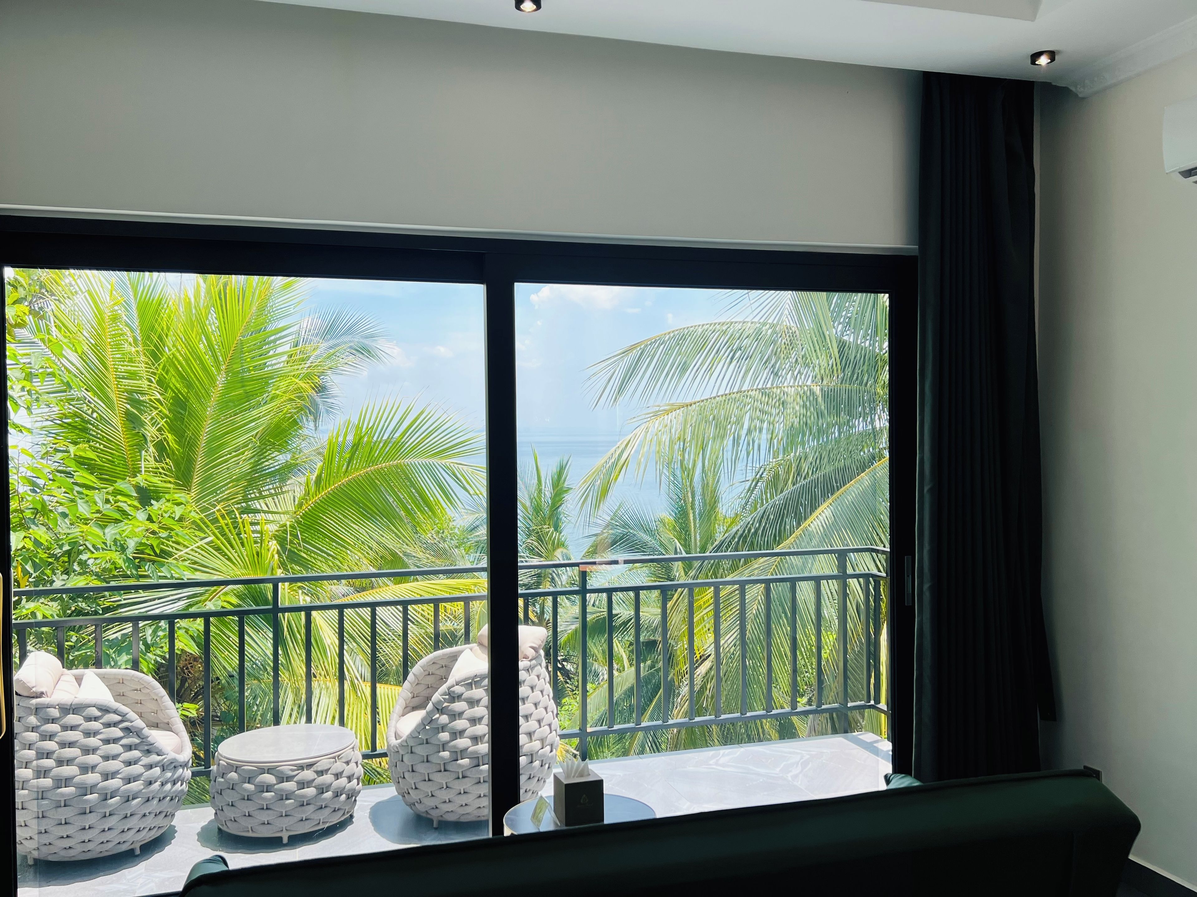 A balcony with a view of palm trees and the sea, furnished with white woven chairs and ottomans.