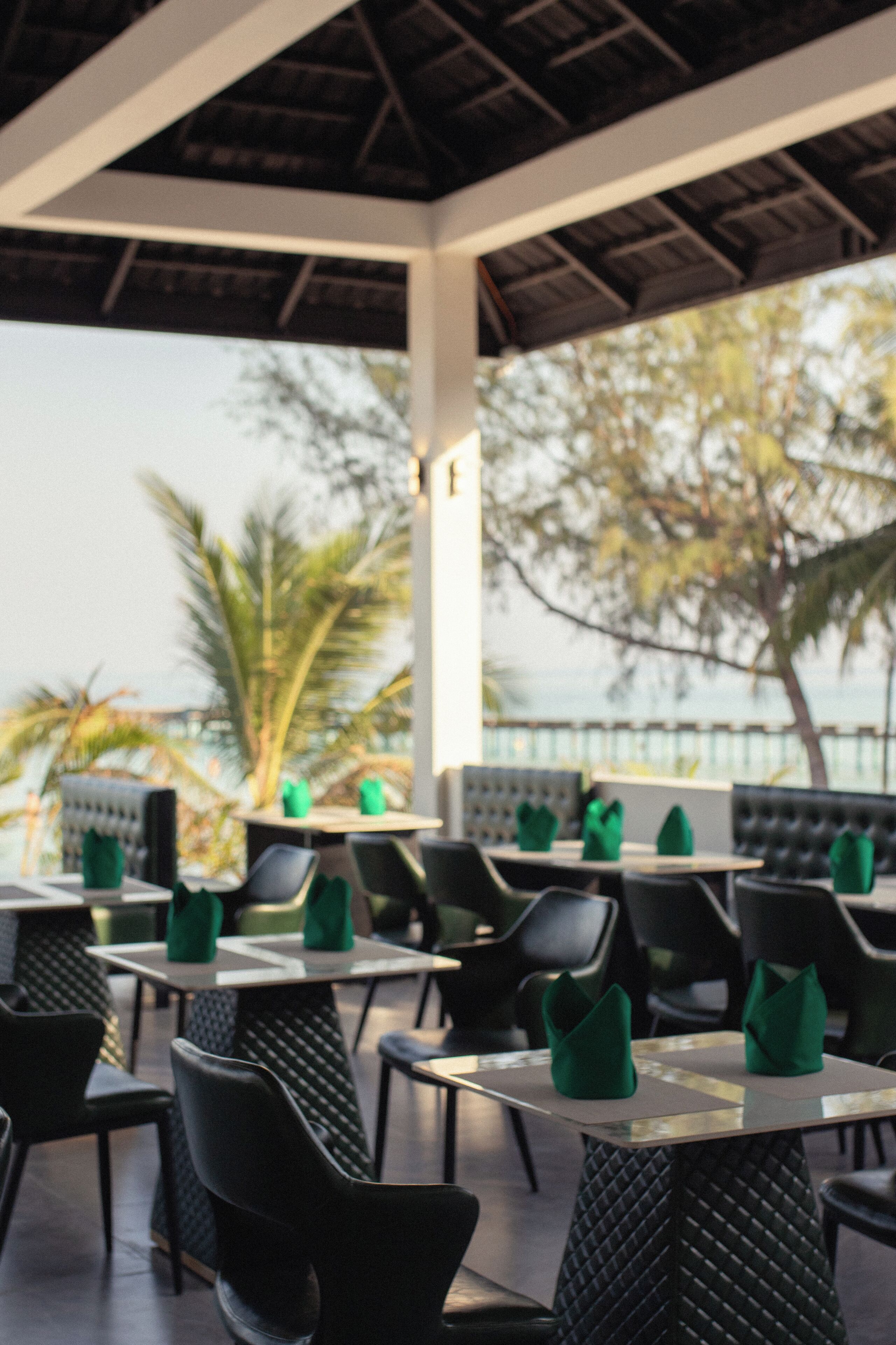 An outdoor restaurant with tables and chairs, a view of the sea, and palm trees.