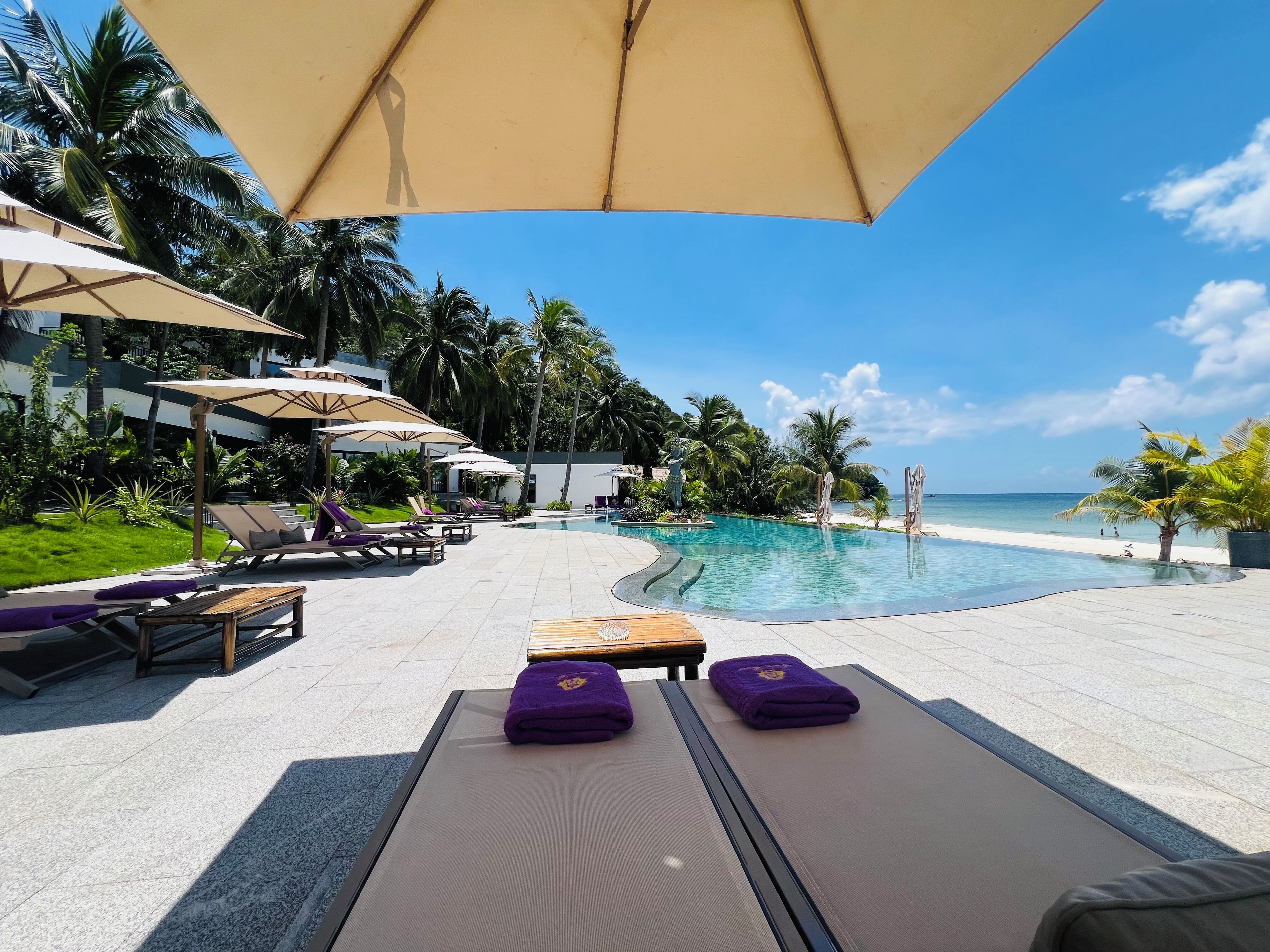 A poolside area with lounge chairs and umbrellas, a clear view of the ocean, and a tropical garden setting.