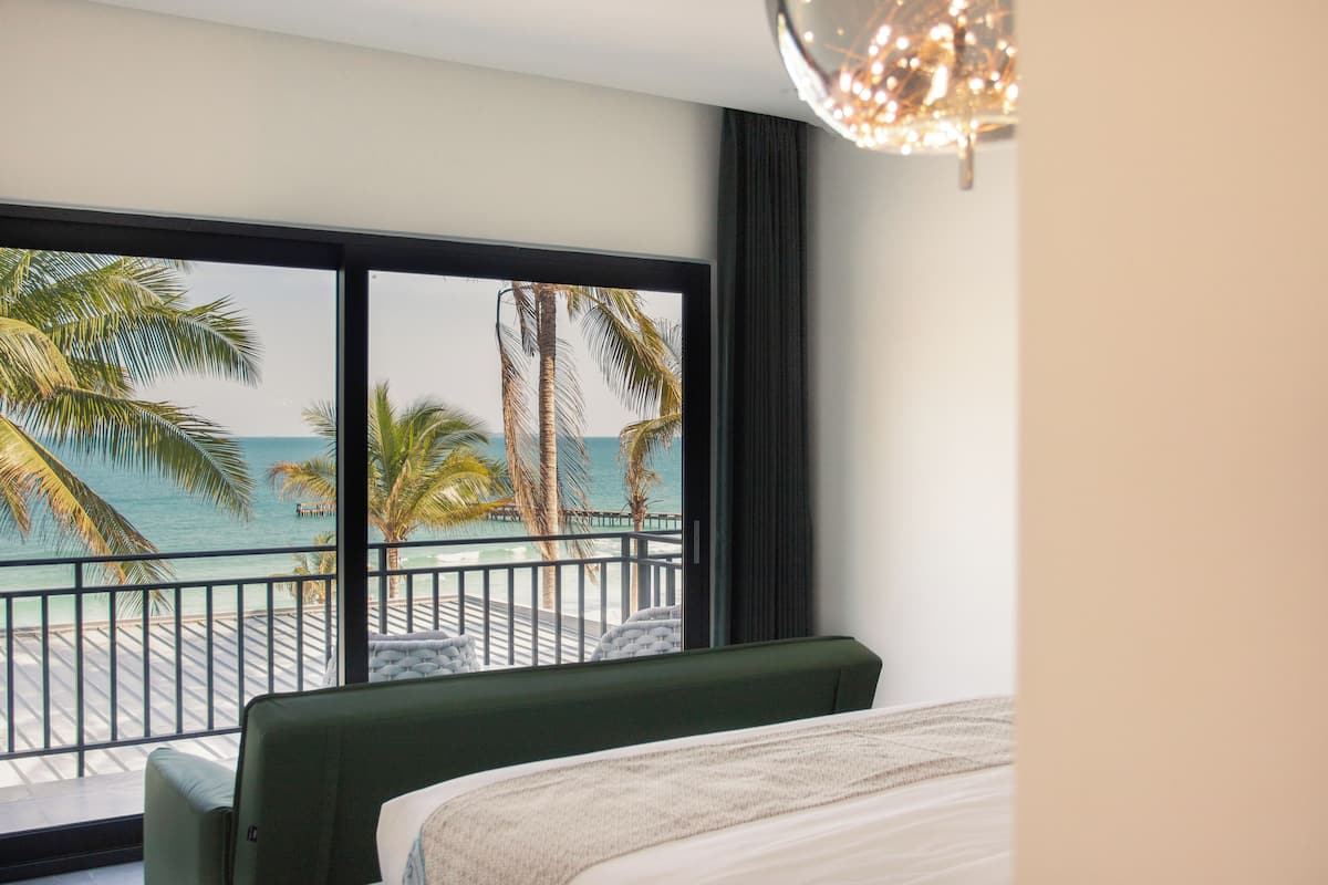 A hotel room with a large window overlooking a beach and ocean, a bed, and a balcony with a railing.