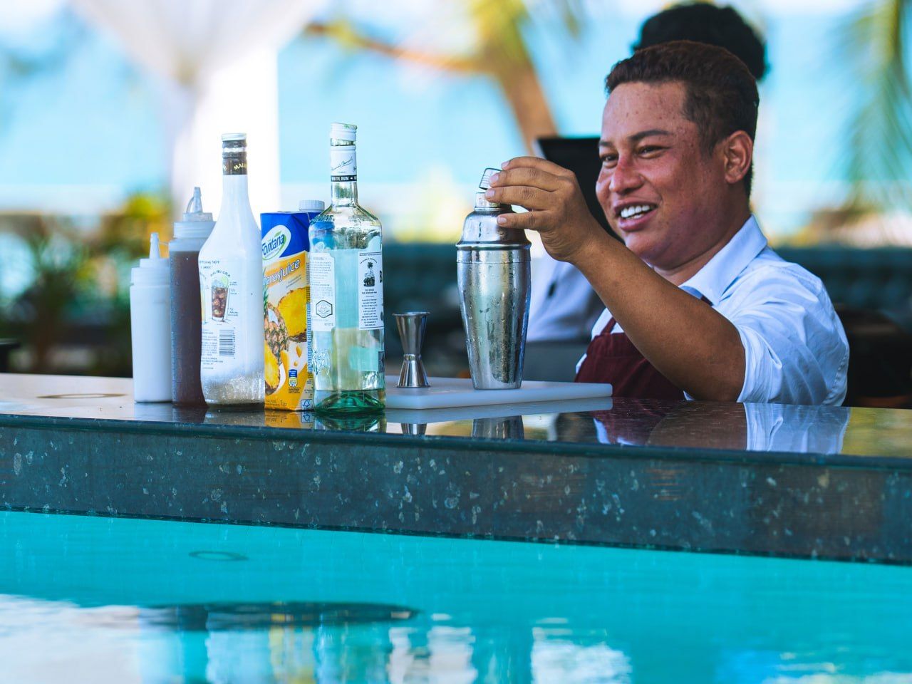 Un cantinero preparando una bebida en un bar junto a la piscina, con varias botellas y un shaker a la vista.