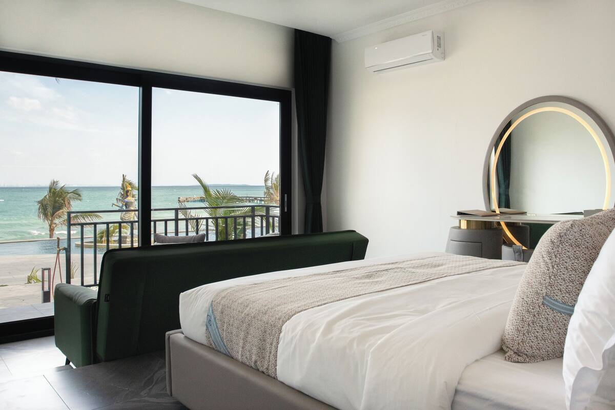 A bedroom with a large window overlooking the ocean, a bed with white bedding, a green upholstered chair, and a desk with a mirror.