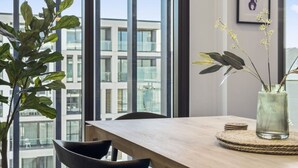 Un interior moderno con una mesa de madera, sillas, un florero con flores y vistas a un edificio a través de grandes ventanales.