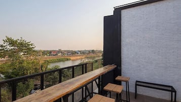 Un balcón con una mesa de madera y taburetes, con vista a un río y edificios al fondo.