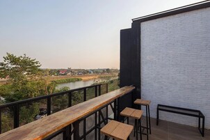 Un balcón con una mesa de madera y taburetes, con vista a un río y edificios al fondo.