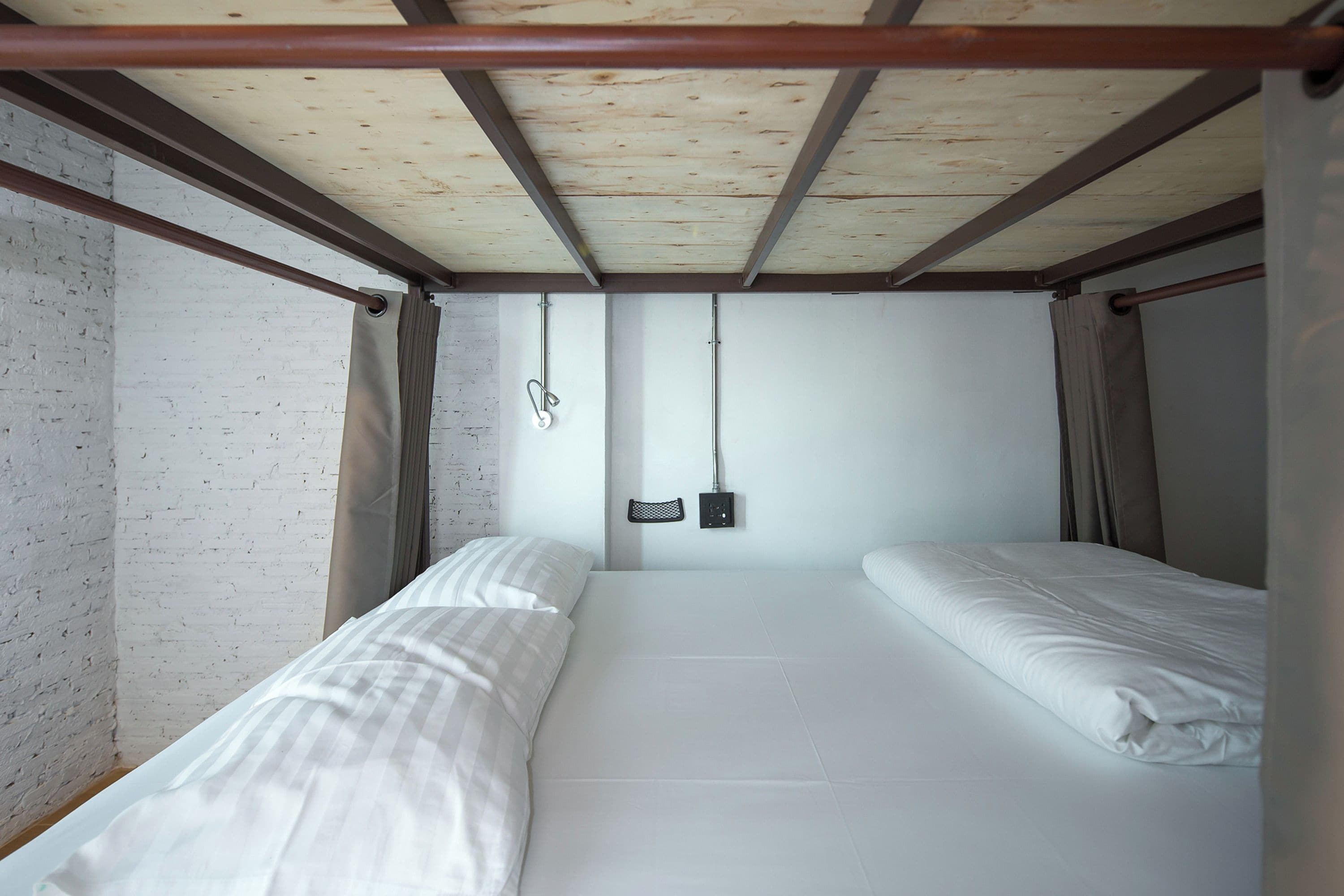 A small, single-bed room with a wooden ceiling and white walls. There are two beds with white linens, a small shelf with a lamp, and a wall-mounted light.