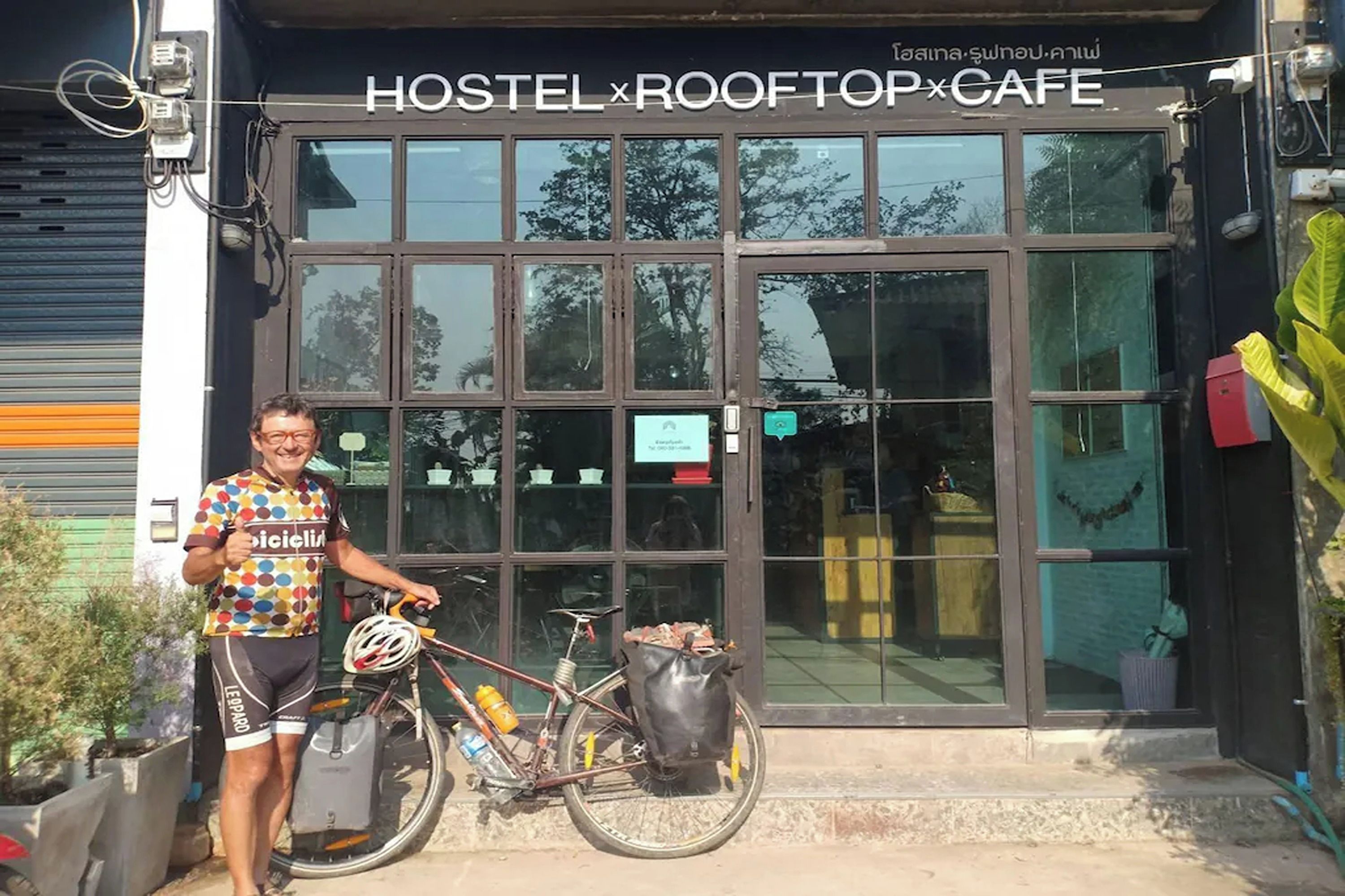 Une entrée d’auberge de jeunesse qui donne sur un toit-terrasse et un café, avec une personne tenant un sac et un vélo.