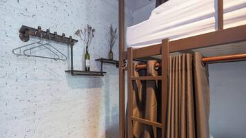 Una cama litera con cortina, un perchero metálico y un estante con una botella y plantas secas.