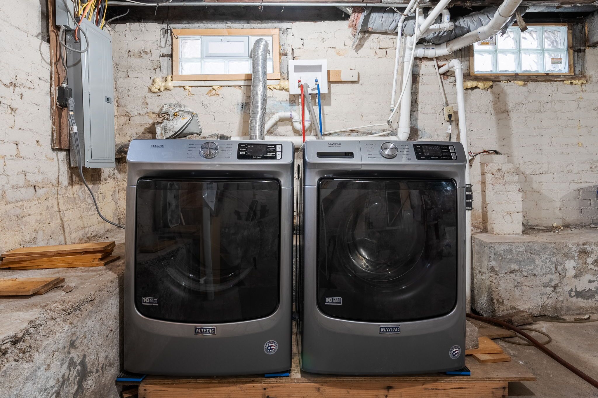 Deux machines à laver Maytag dans une pièce aux murs en briques apparentes, avec des câbles électriques visibles et une fenêtre donnant sur un paysage.