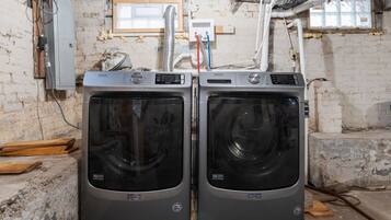 Deux machines à laver Maytag dans une pièce aux murs en briques apparentes, avec des câbles électriques visibles et une fenêtre donnant sur un paysage.