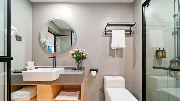 A modern bathroom with a round mirror, white sink, and toilet. There is a towel rack above the toilet and a shelf with folded towels. - Yangshuo Mountain and City Resort  (Guilin)