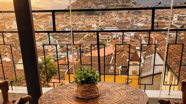 Een ronde houten tafel met een geweven decoratie in het midden, omringd door houten stoelen met gebeeldhouwde details, op een balkon met uitzicht op de stad.