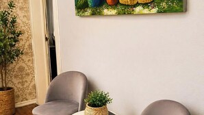 Une pièce décorée d’un tableau représentant une femme entourée de fleurs, de deux fauteuils gris, d’une petite table ronde ornée d’une plante et d’une plante en pot.