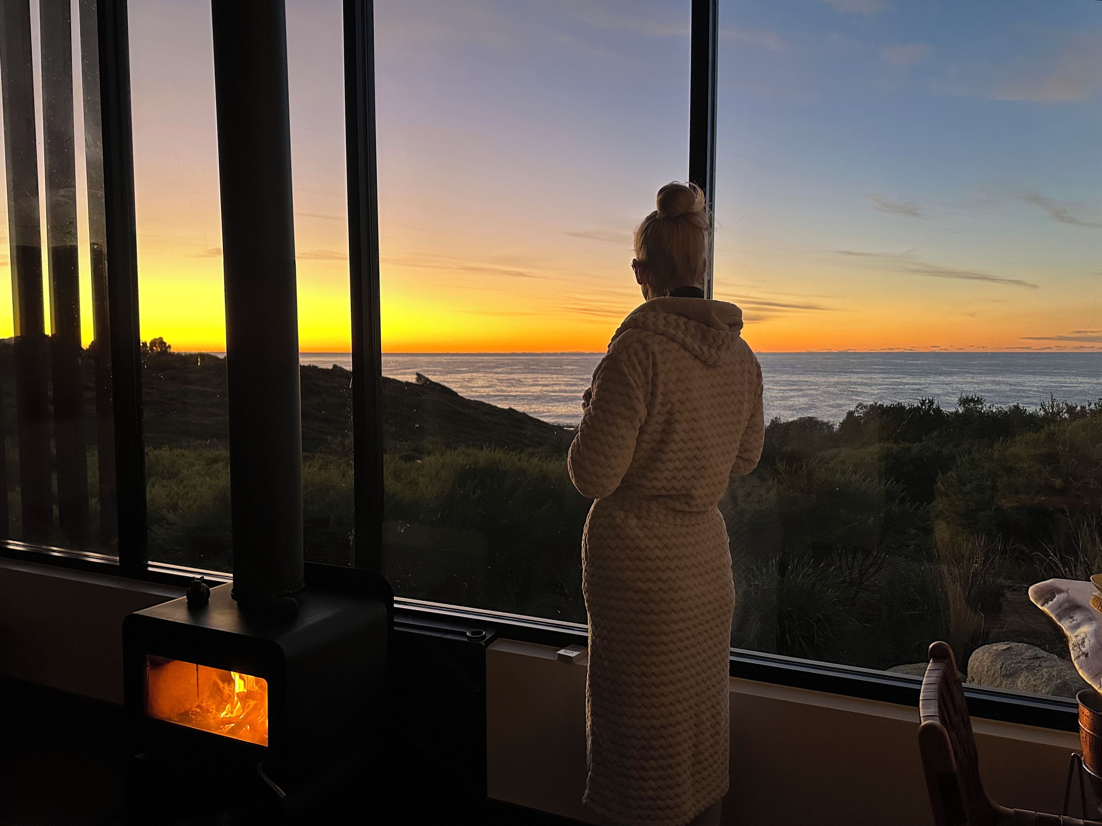 A person in a robe standing by a large window overlooking a sunset, with a fireplace visible in the foreground.