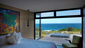 Une chambre avec une grande fenêtre donnant sur l’océan, une peinture abstraite aux couleurs vives accrochée au mur et un oreiller blanc moelleux sur le lit.