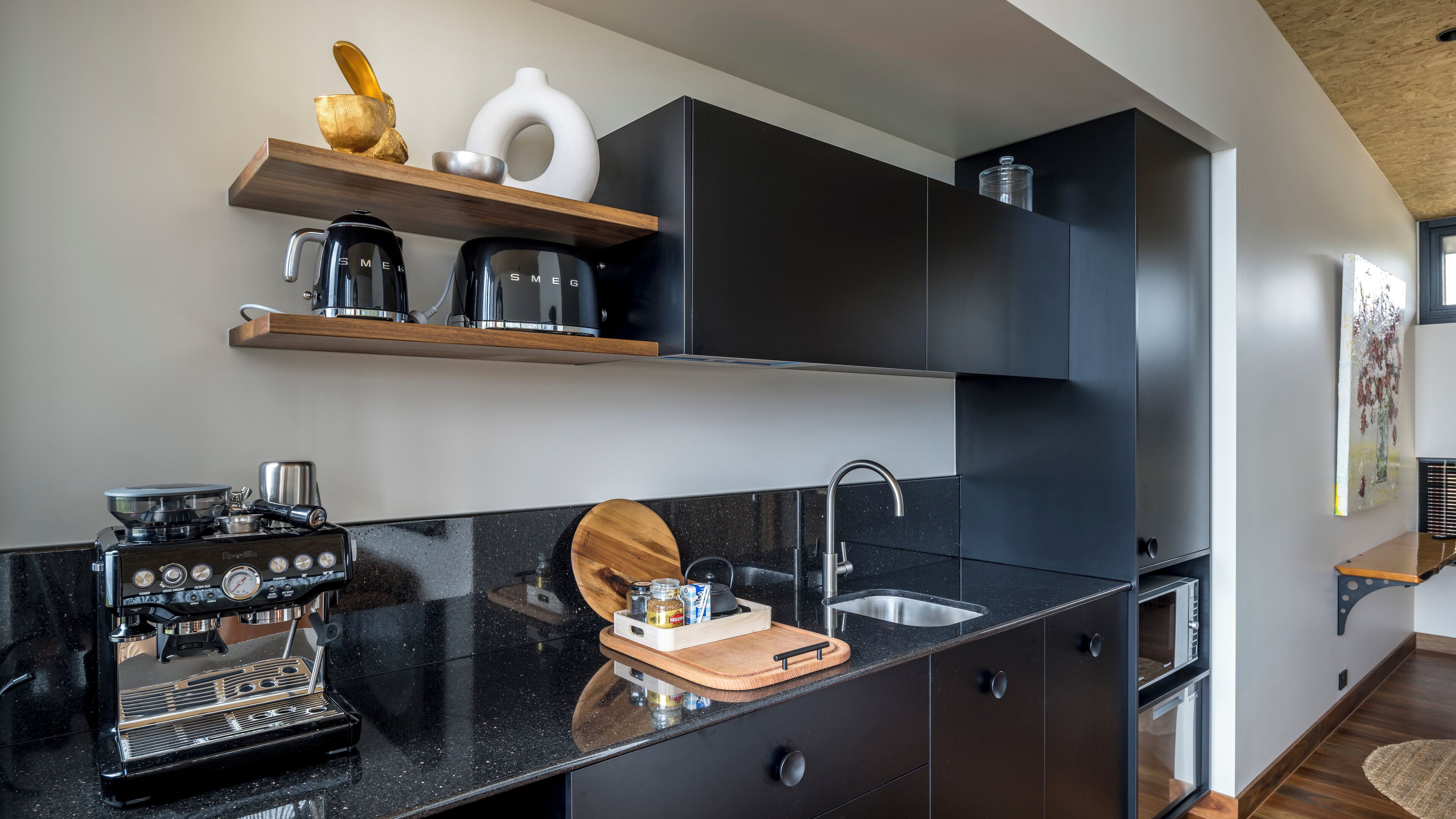 A modern kitchen with a black countertop, a coffee machine, a sink, and wooden shelves above.