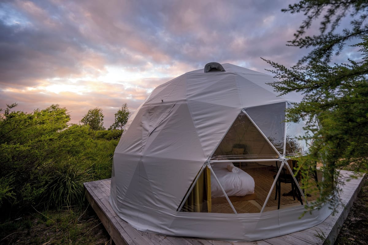 A geodesic dome tent with a wooden deck, surrounded by greenery.
