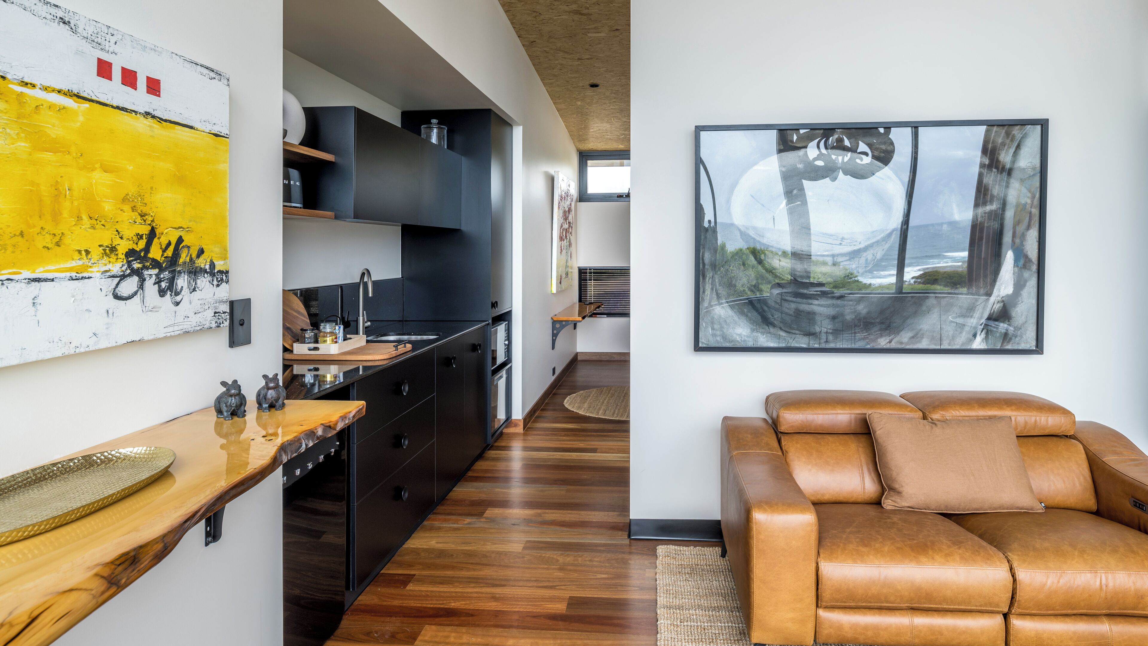 A modern kitchen with dark cabinetry, a wooden countertop, and a view of the living area with a leather sofa and a large abstract painting.