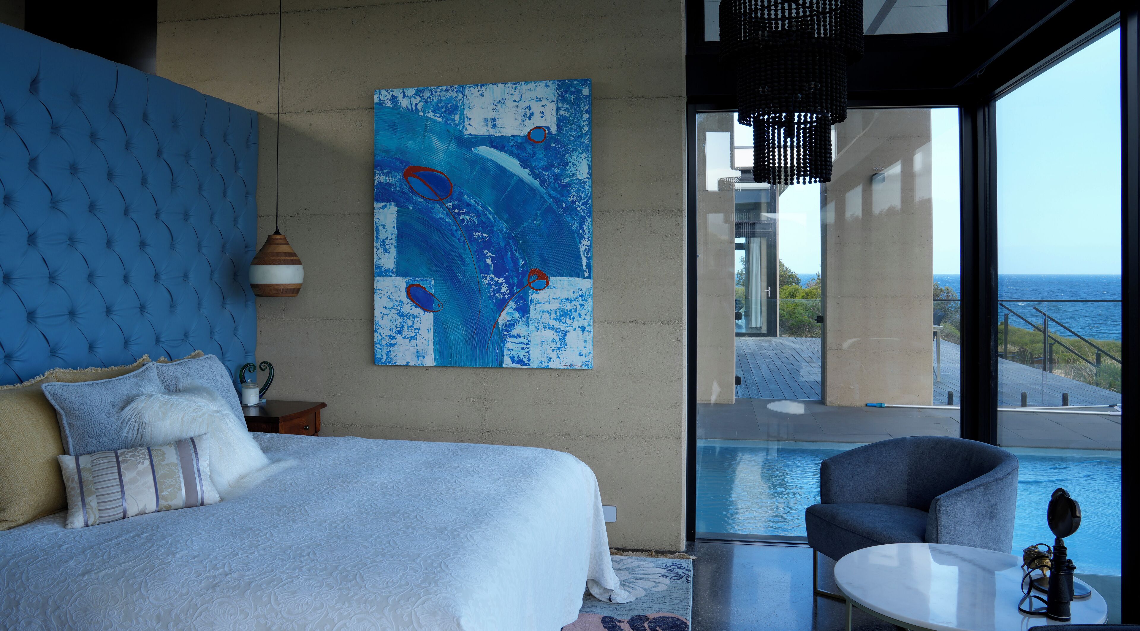 A bedroom with a large bed, a painting, a view of the ocean, and a pool area.