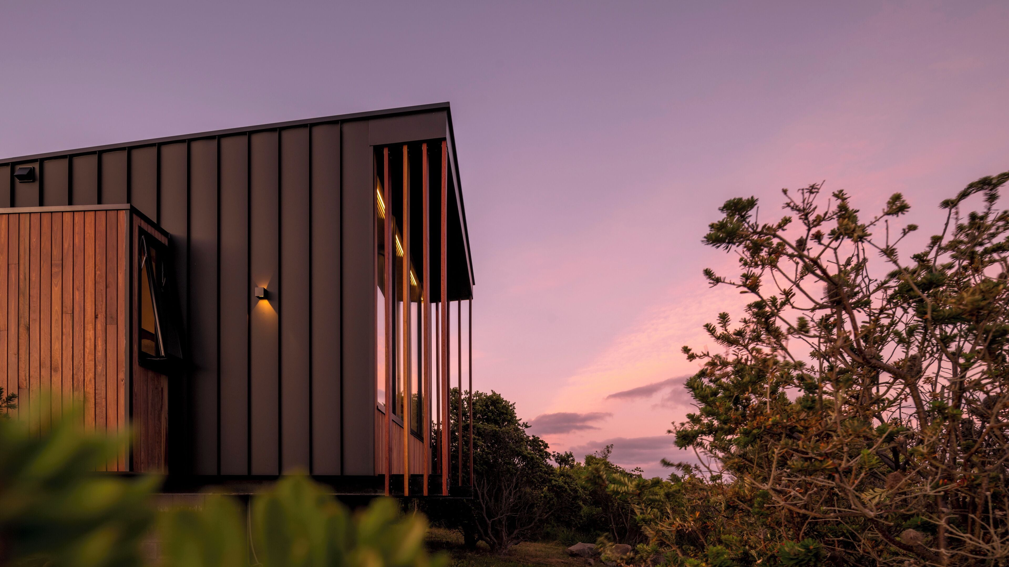A modern building with a wooden facade and a large window, surrounded by dense vegetation.