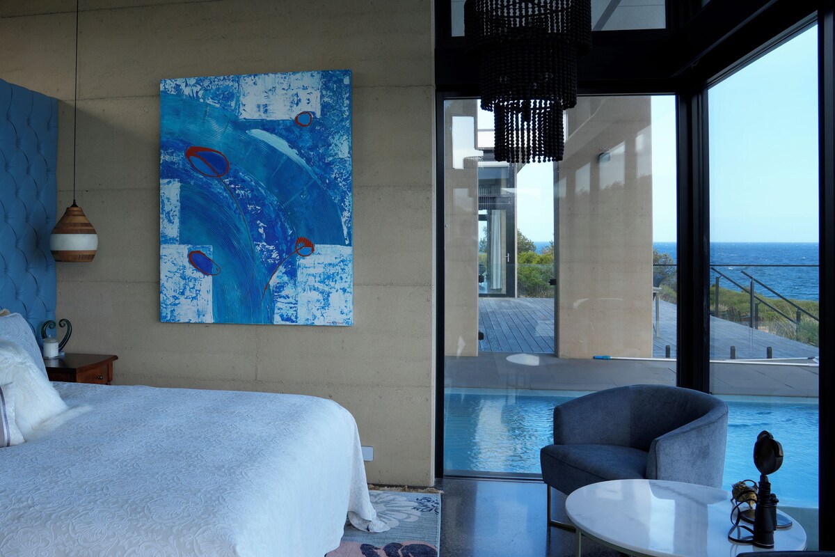 A bedroom with a large bed, a painting, a view of the ocean, and a pool area.