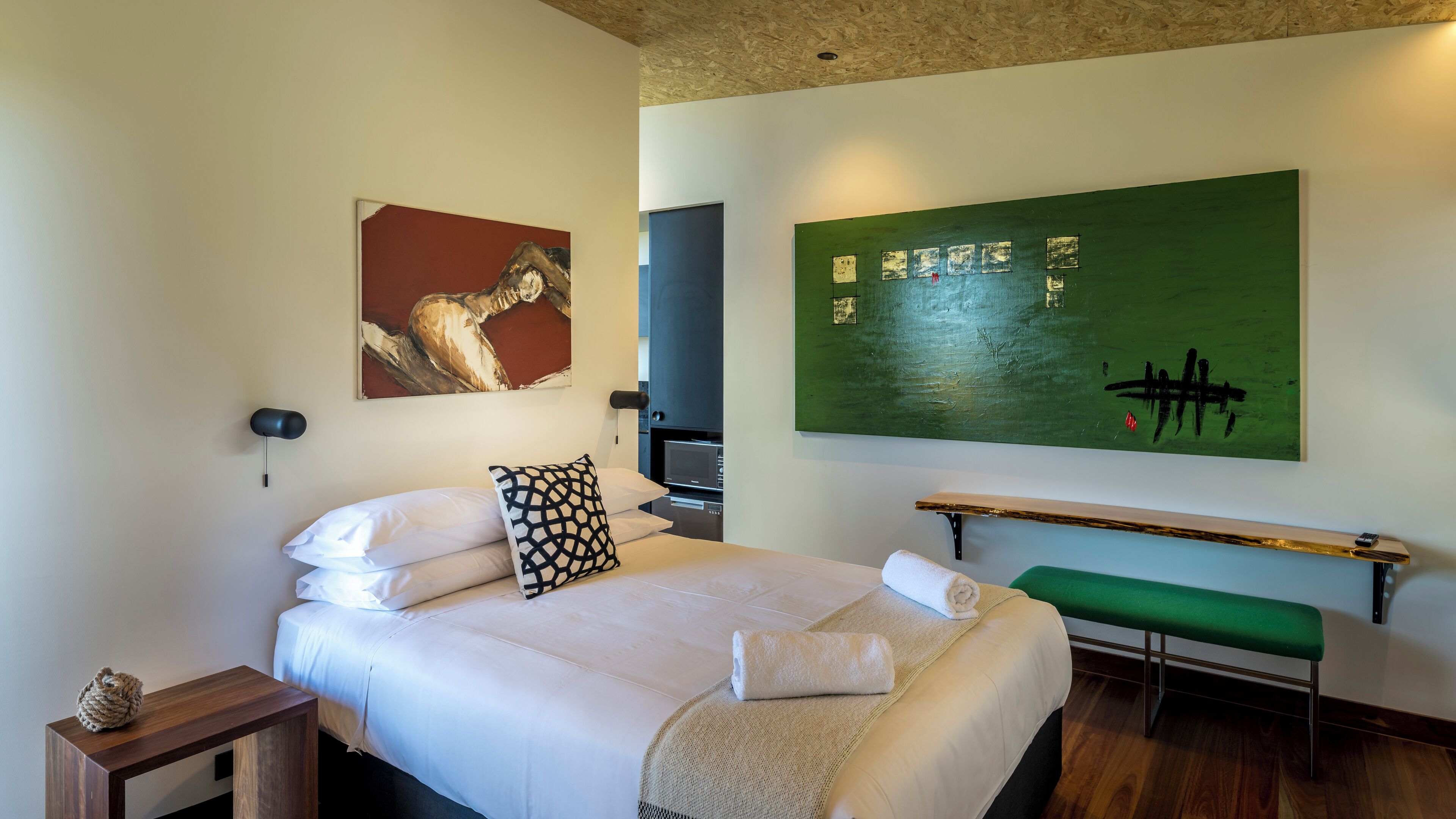 A hotel room with a bed, a green bench, a wall-mounted shelf, and artwork.