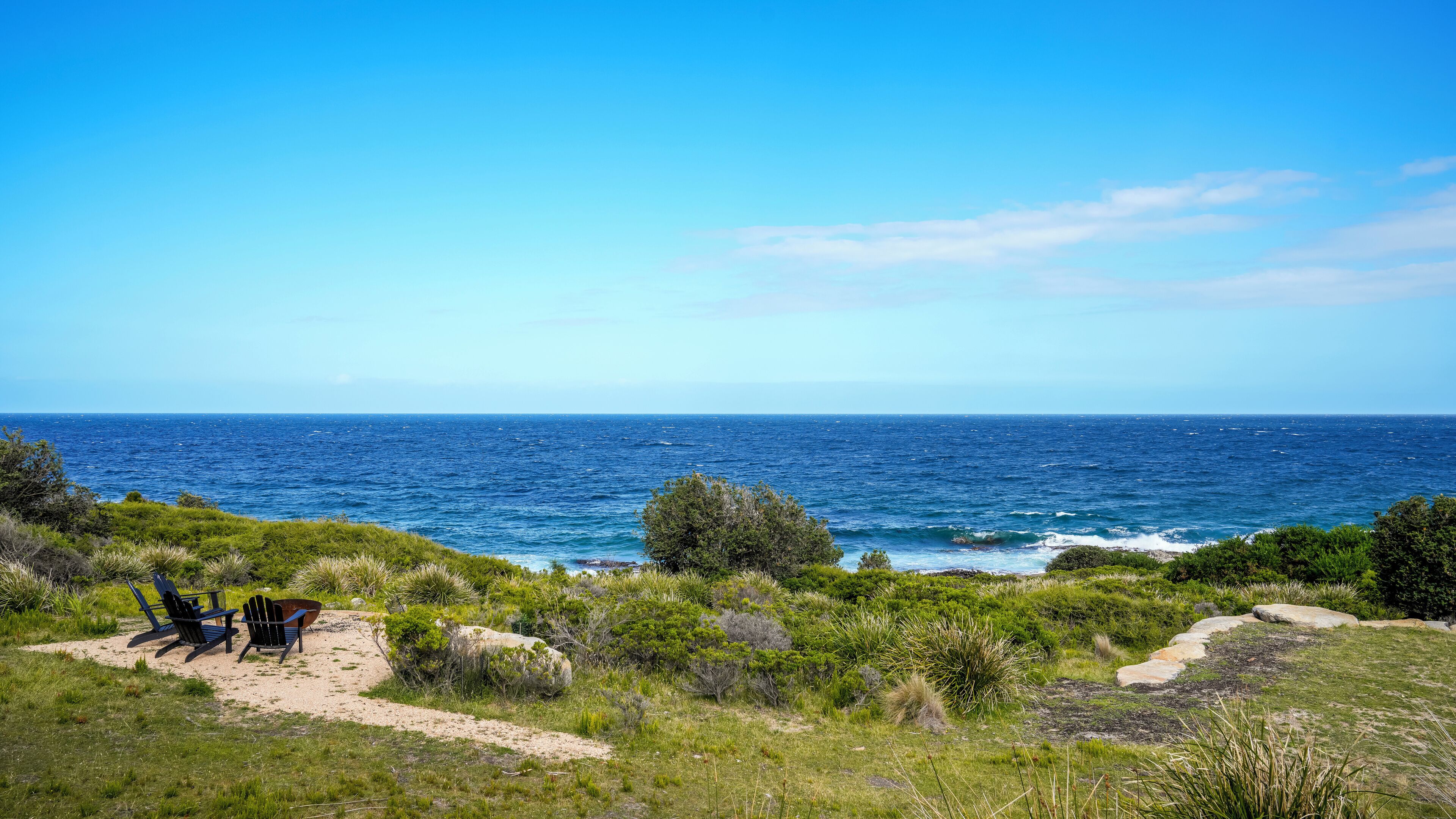 Paesaggio costiero con vista sull'oceano, un'area sabbiosa con alcune sedie e vegetazione.