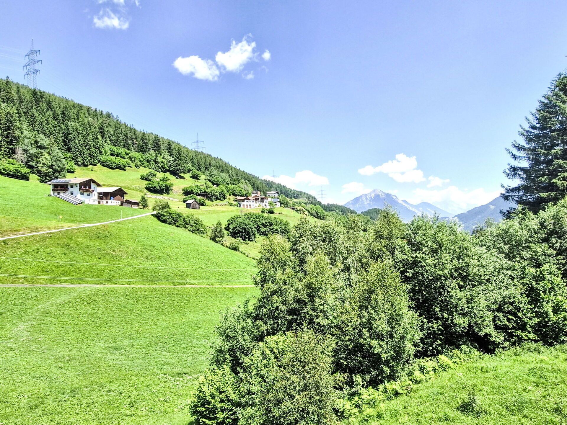 Un paisaje rural con una casa en una ladera, rodeada de abundante vegetación y montañas al fondo.