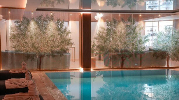 Une piscine intérieure moderne aux parois en verre transparent, offrant une vue sur des plantes d’intérieur et le paysage urbain.