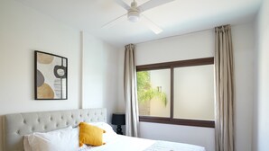 Un lit bien fait, recouvert d’une courtepointe matelassée, avec un seul oreiller jaune et des serviettes enroulées posées sur le côté. On aperçoit un ventilateur de plafond au-dessus du lit.