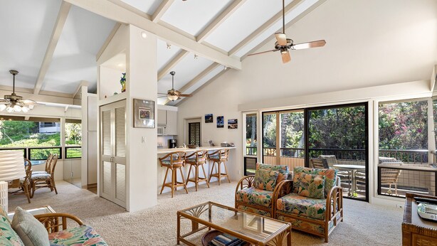 A spacious living area with a high ceiling, a dining area with bar stools, a sofa with floral upholstery, and large windows offering a view of the outdoors.