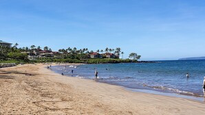 A beach with clear skies, people swimming and walking along the shore, and buildings with red roofs in the background.