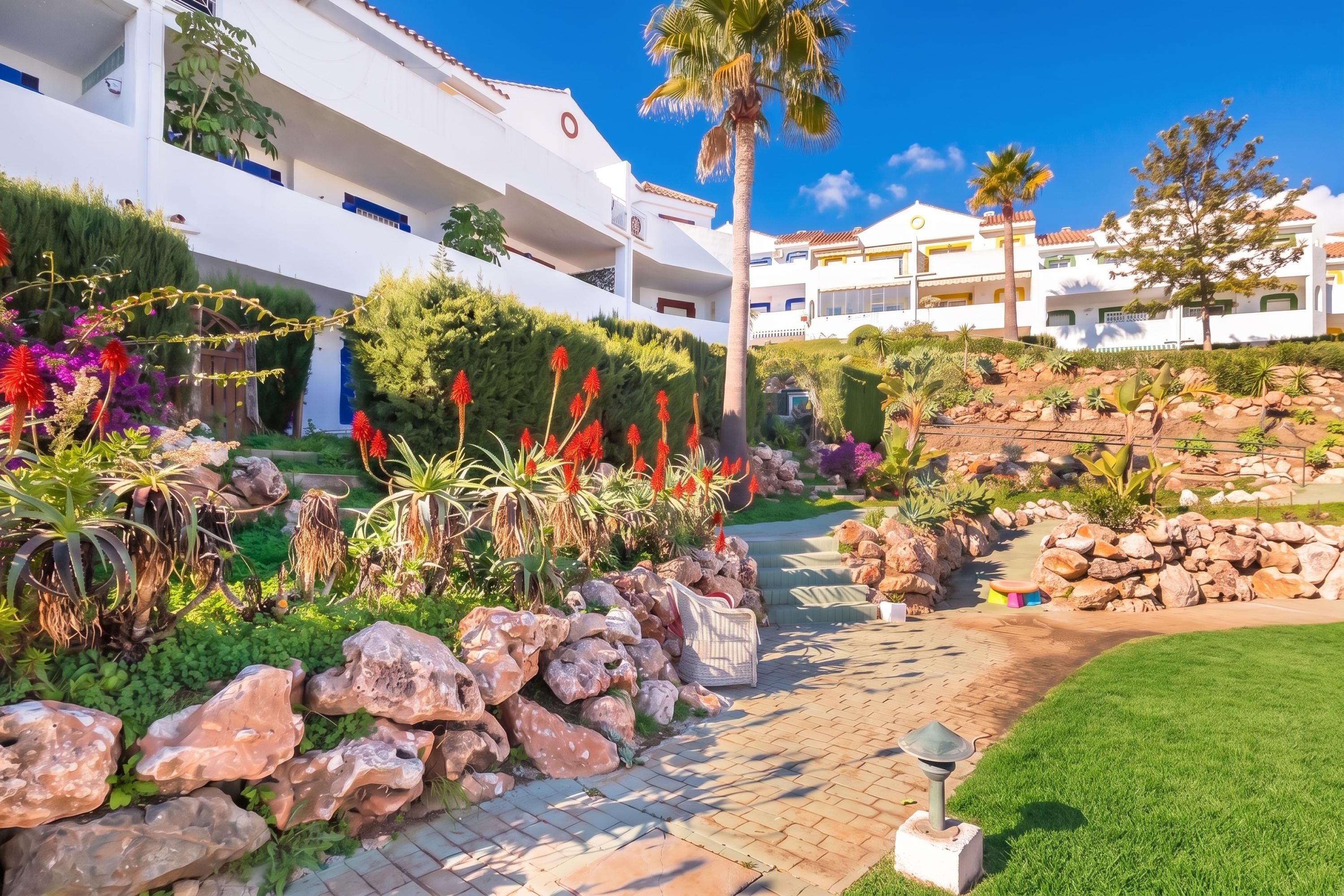 A pathway leading to a building with a white facade and balconies, surrounded by landscaped gardens with rocks and plants.