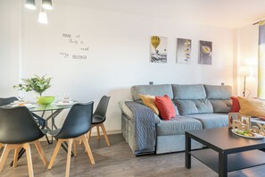 A modern living room with a grey sofa, black chairs, a glass dining table, and a green bowl. There are wall art pieces and a sign that reads "TIME with Family is worth every SECOND".