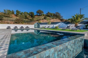 Ein rechteckiger Pool mit Fliesenrand, umgeben von Liegestühlen und einer kleinen Grünanlage.