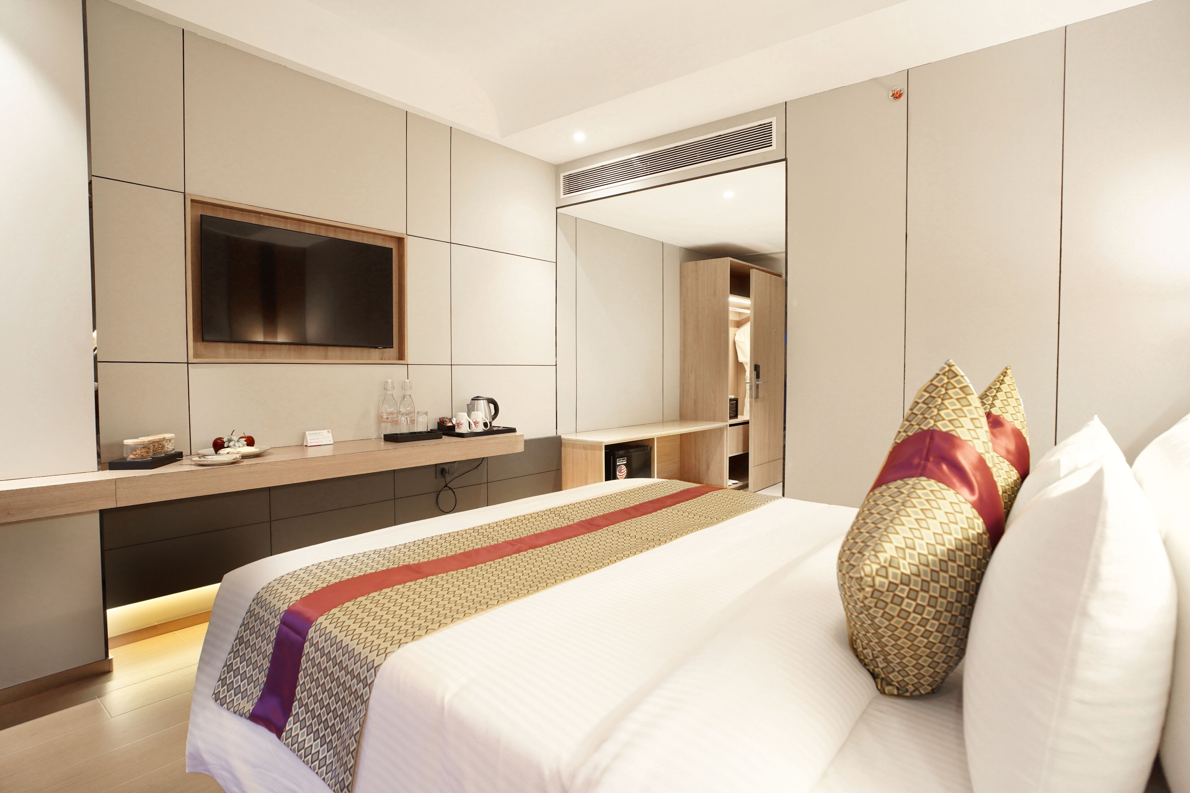 Une chambre d’hôtel moderne dotée d’un grand lit, d’un téléviseur à écran plat fixé au mur, d’une armoire intégrée et d’une table de chevet avec une bouilloire et des tasses.