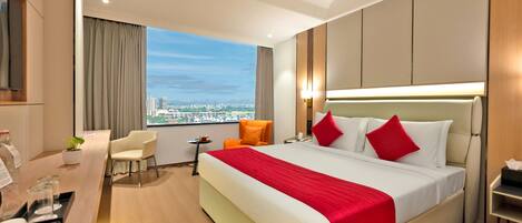 A hotel room with a large bed, a desk with a chair, a small table with a fruit plate, and a view of the cityscape.