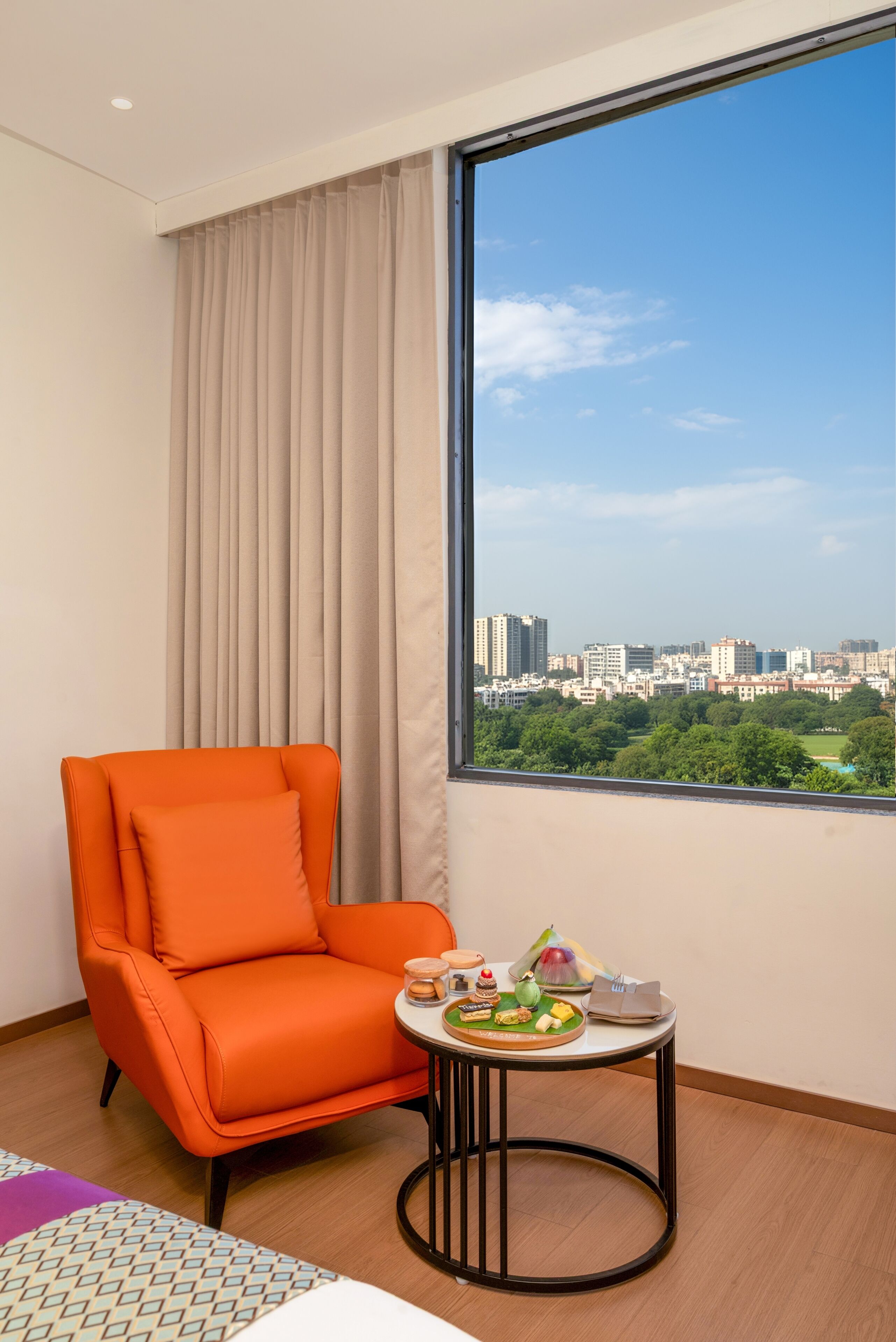 Habitación de hotel con un ventanal grande, una silla naranja individual, una pequeña mesa redonda con una bandeja de comida y vista a la ciudad.