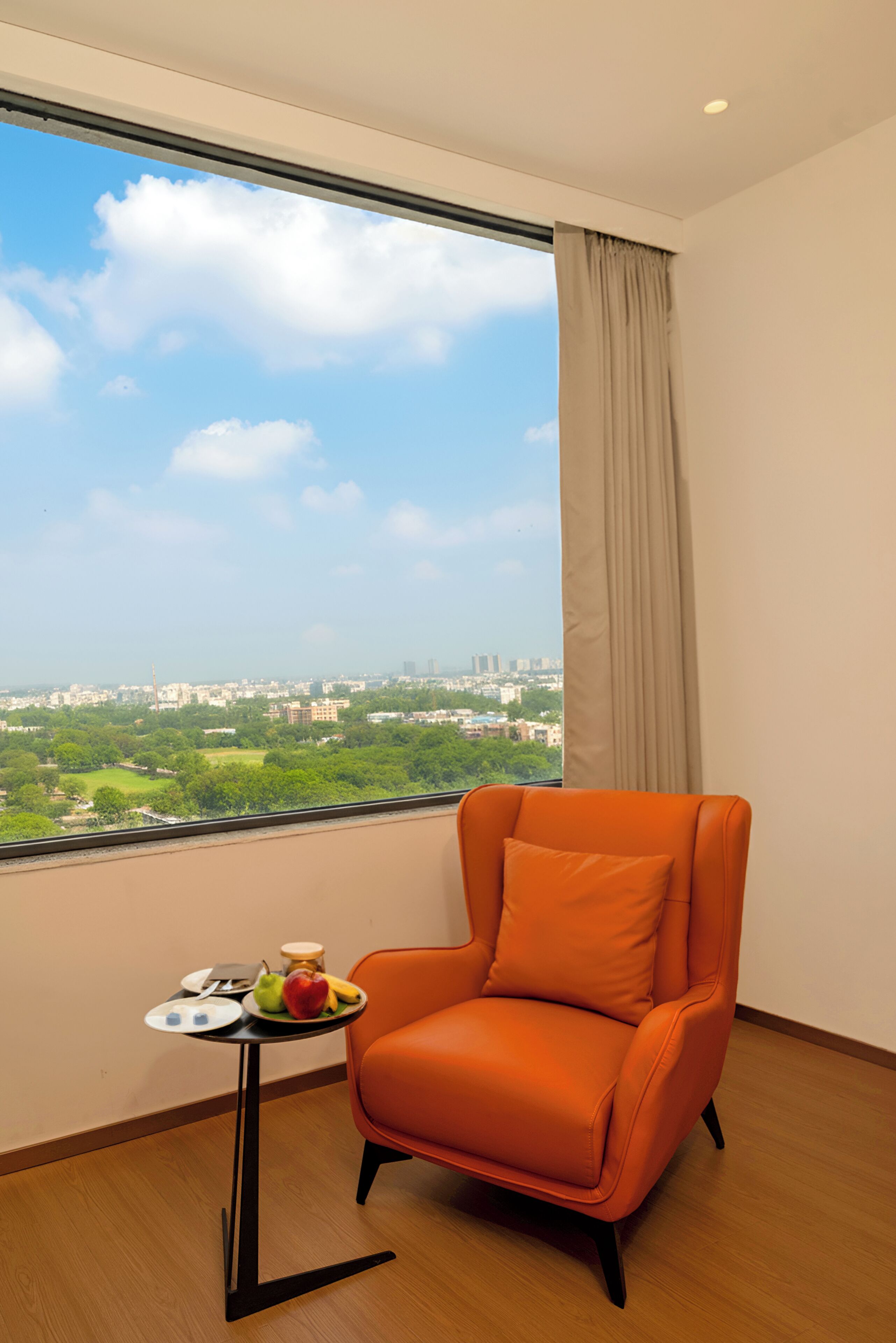 A room with a large window, a single orange chair, and a small table with fruits and a jar.