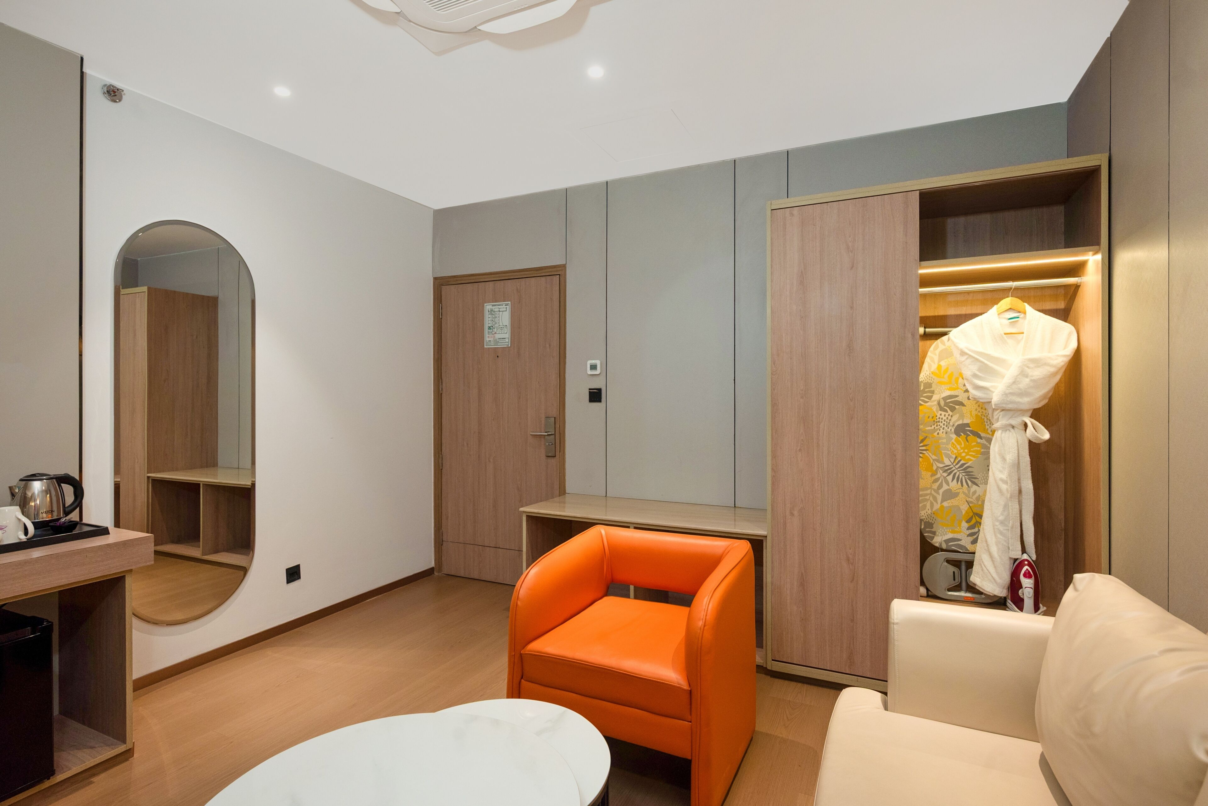 A modern hotel room with a wooden wardrobe, a small table with a kettle, a bright orange chair, and a white sofa.
