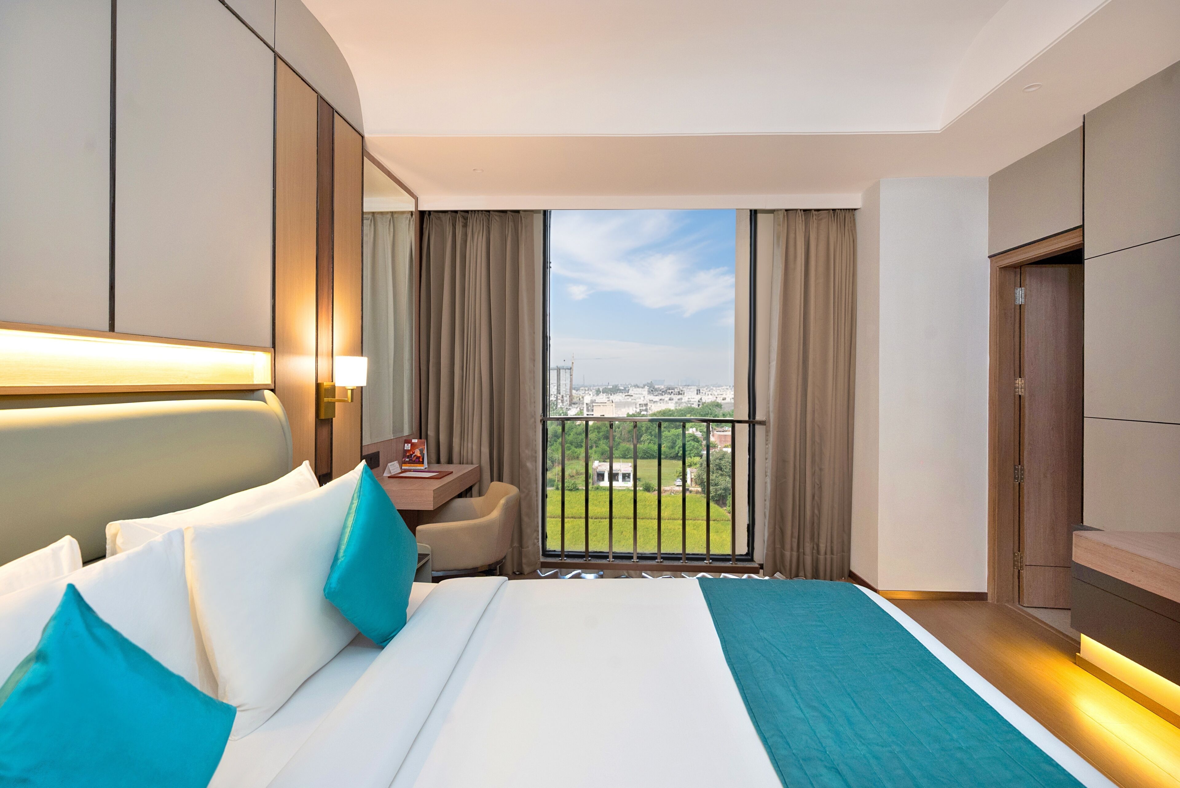 Habitación de hotel con una cama grande, un escritorio, una silla y vistas a un paisaje urbano desde un balcón.