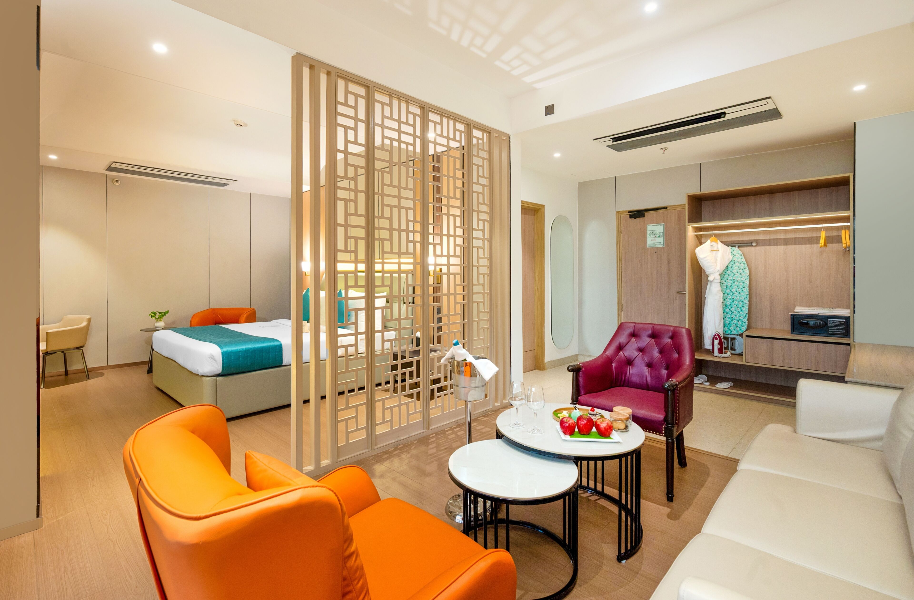 Habitación de hotel con cama, sillones naranja, sofá blanco, mesa de comedor con una bandeja de frutas y armario con ropa colgada.